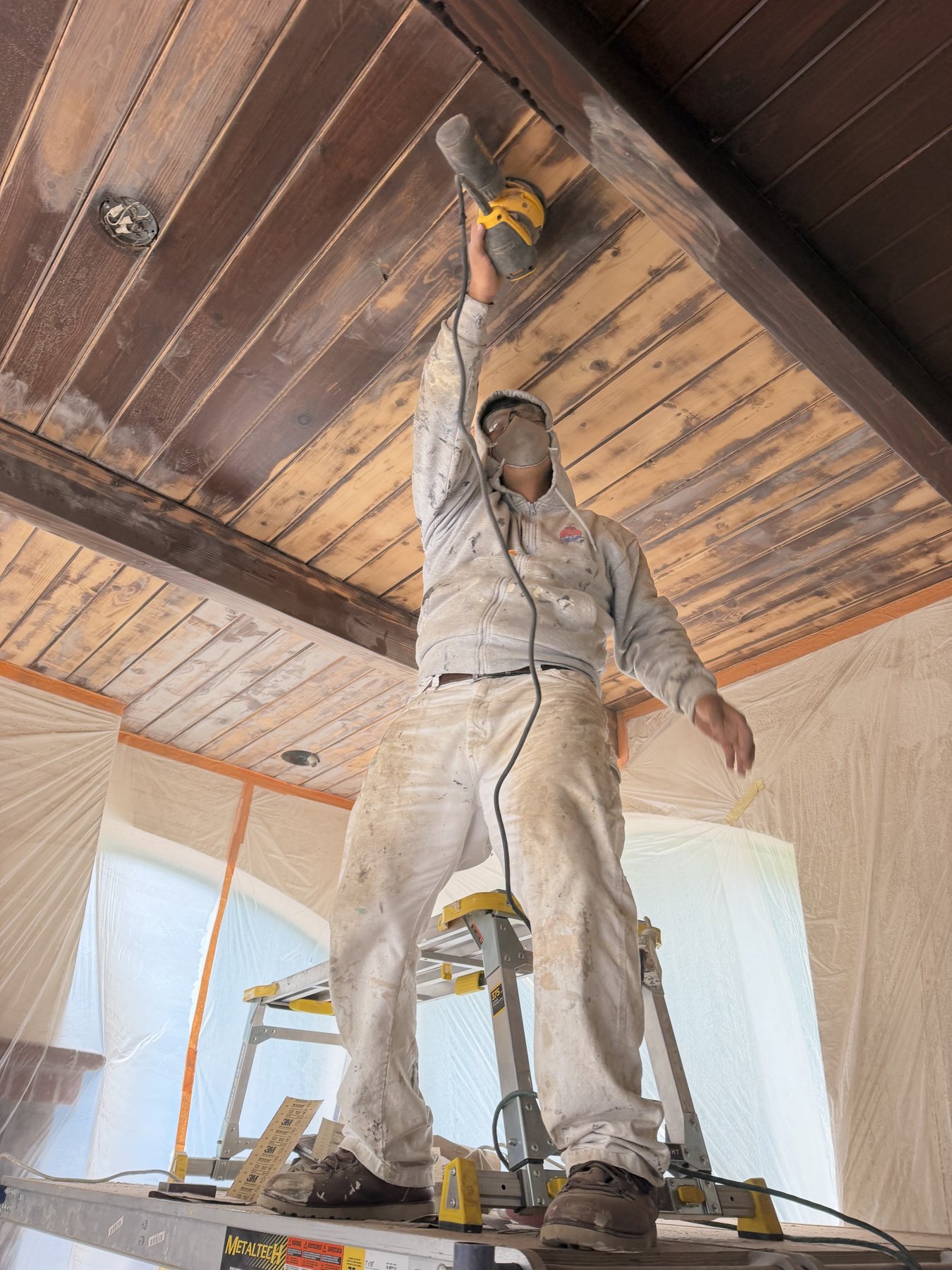 Painter sanding a wood ceiling with the surrounding room masked and protected